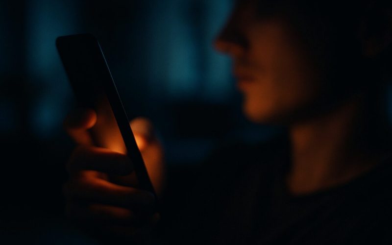 A close, slightly abstract night scene of a smartphone held in one hand, its face turned mostly away from the camera so only a soft featureless amber glow spills out across the fingers and jawline just outside focus. Dark blurred living room behind, deep blues against warm highlights, shallow depth of field. Cinematic editorial photography with a quiet, uneasy mood.